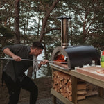 A man using a black wood-fired pizza oven outdoors in a wooded setting, with stacked firewood and a wooden work surface beside the oven.